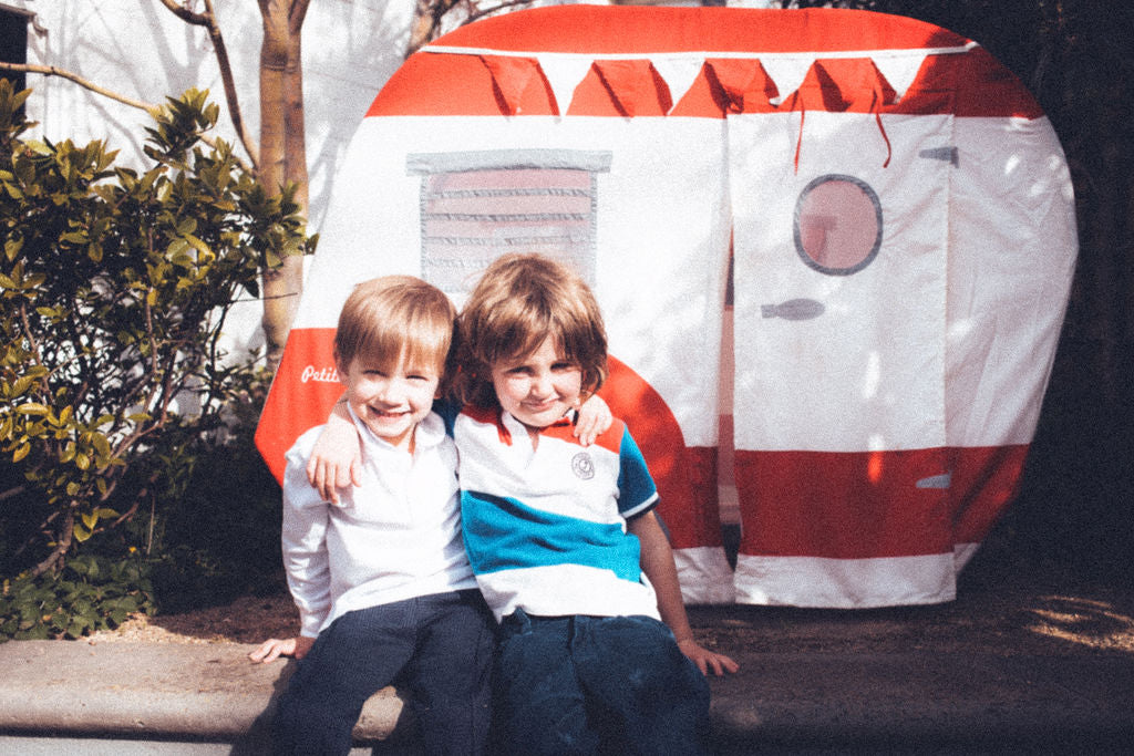 Two children sitting in front of aPetite Maison Play Vintage Camper Cubby House outdoors.